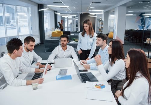 Guy on the left side talking and colleagues is listening to him. Group of young freelancers in the office have conversation and smiling.