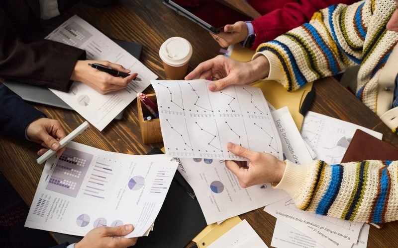 Group of young adult and middle aged Caucasian and Black hands analyzing business charts and graphs at table, holding printed documents with data, discussing financial strategy, coffee cup visible